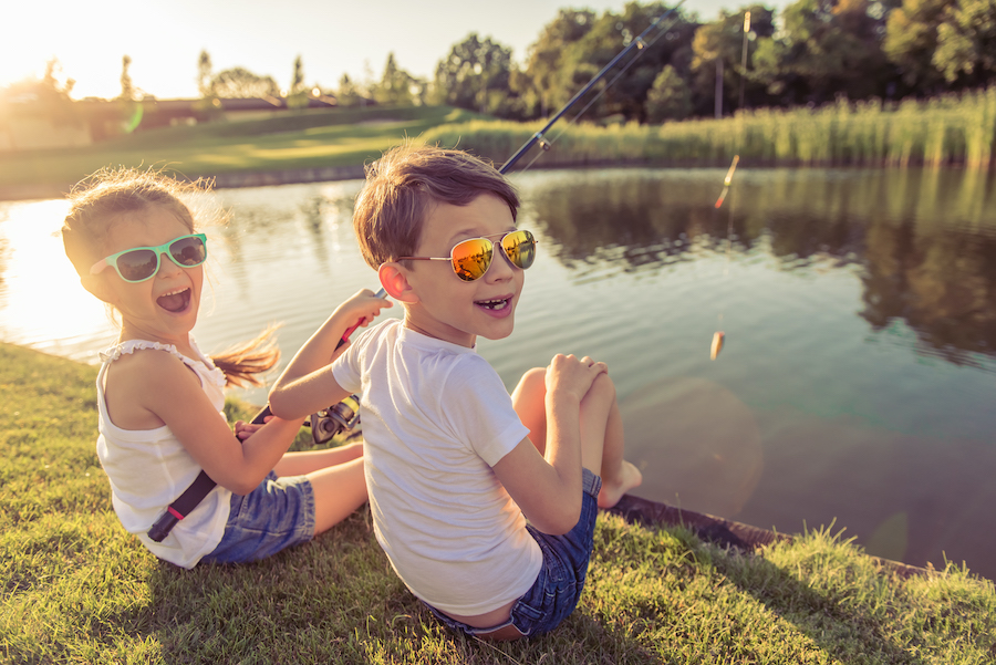 Smiling young boy and girl wear sunglasses as they sit along the water fishing in the summer