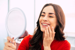 Brunette woman in a red blouse smiles while looking at her teeth in a hand mirror